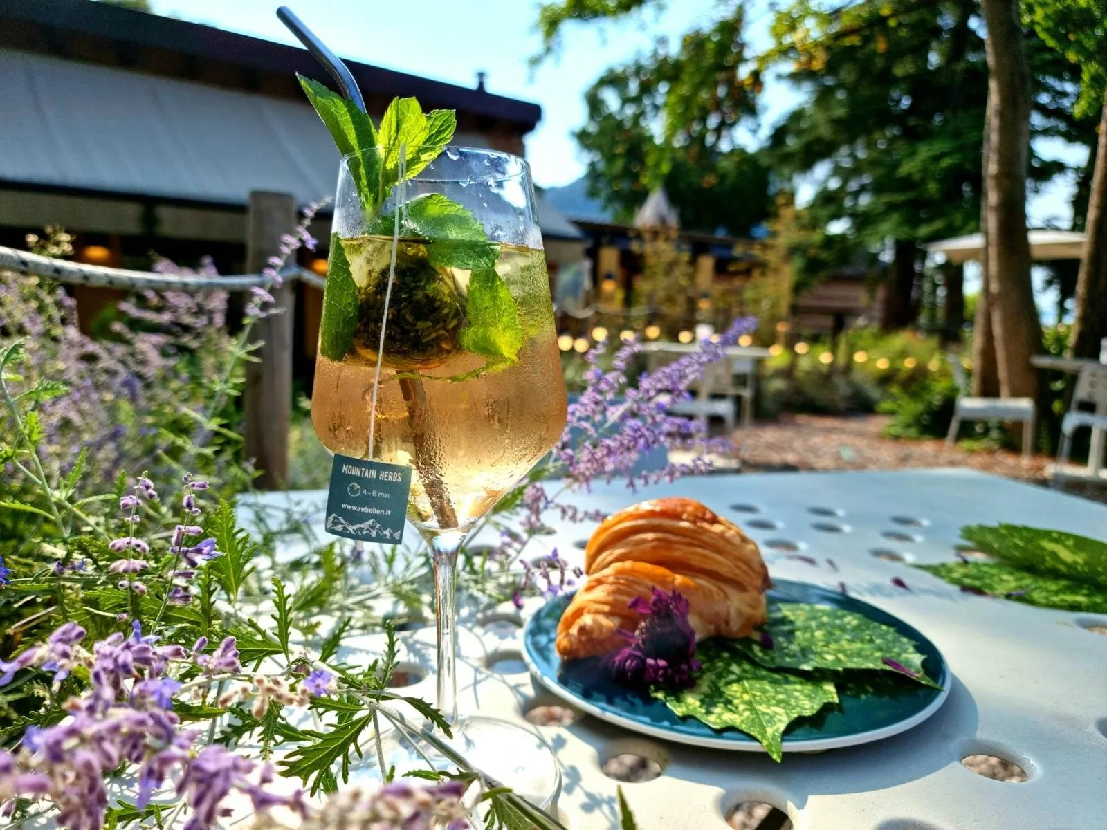 Un cocktail con menta e lavanda accanto a un cornetto, su un tavolo del giardino nella luce calda del tardo pomeriggio