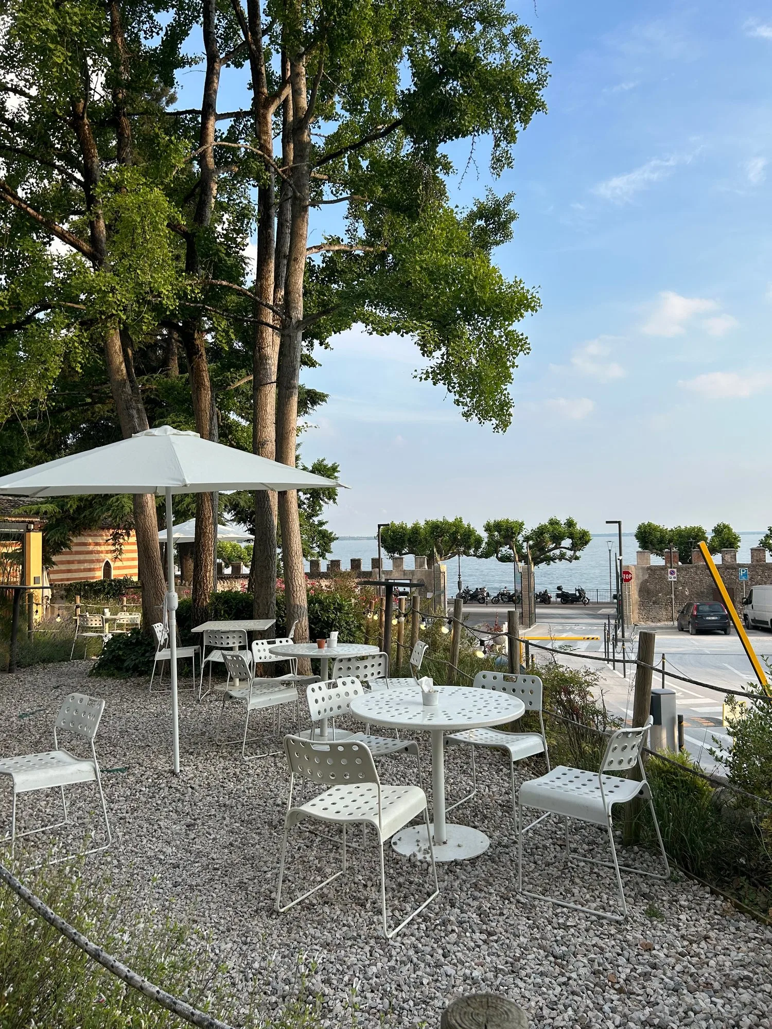 Tavolini bianchi tra gli alberi del giardino Hortus, con uno scorcio del lago di Garda sullo sfondo